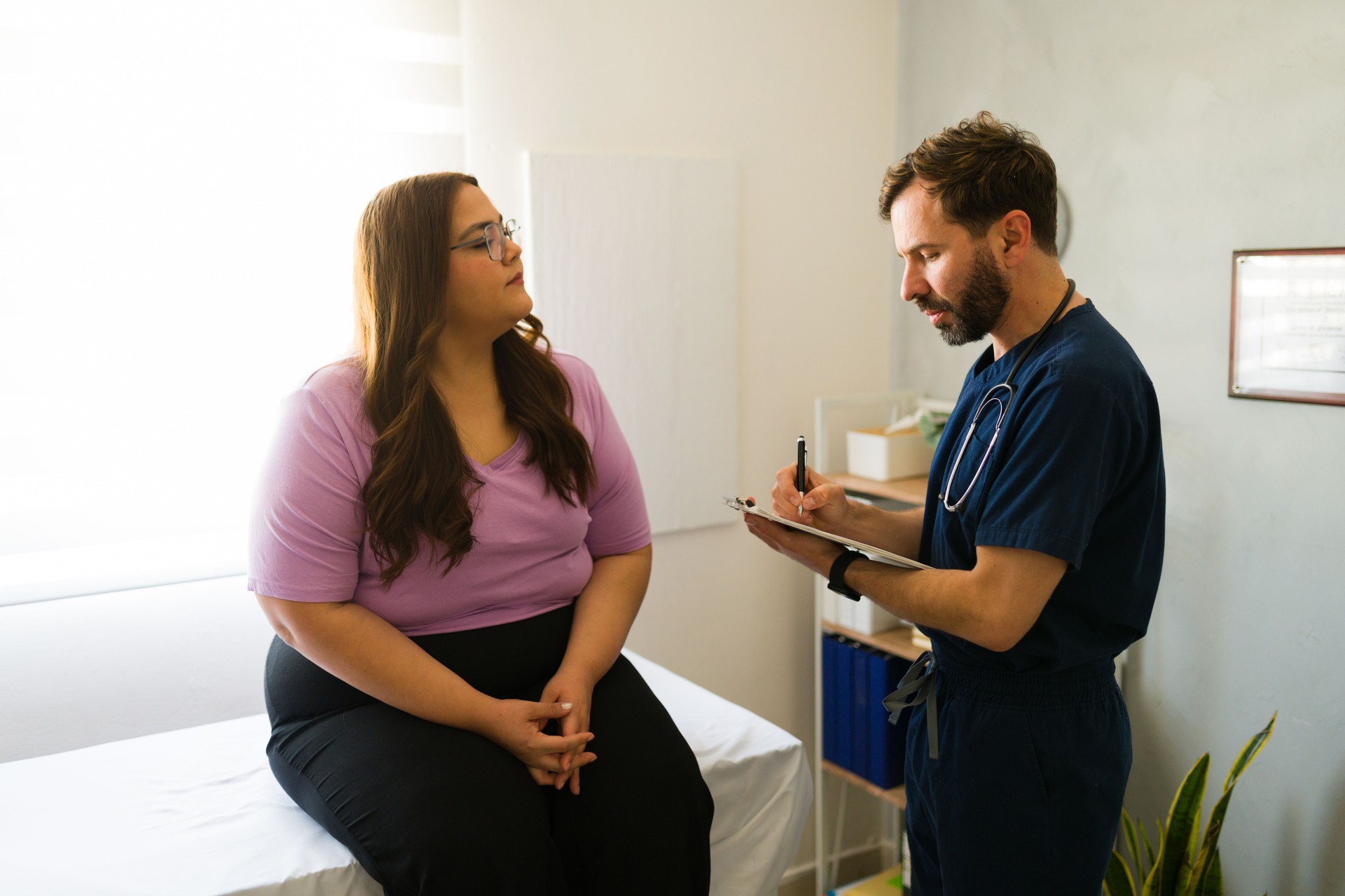 Paciente femenina de talla grande conversando con un profesional de la salud, tomando notas durante la consulta médica en un entorno clínico